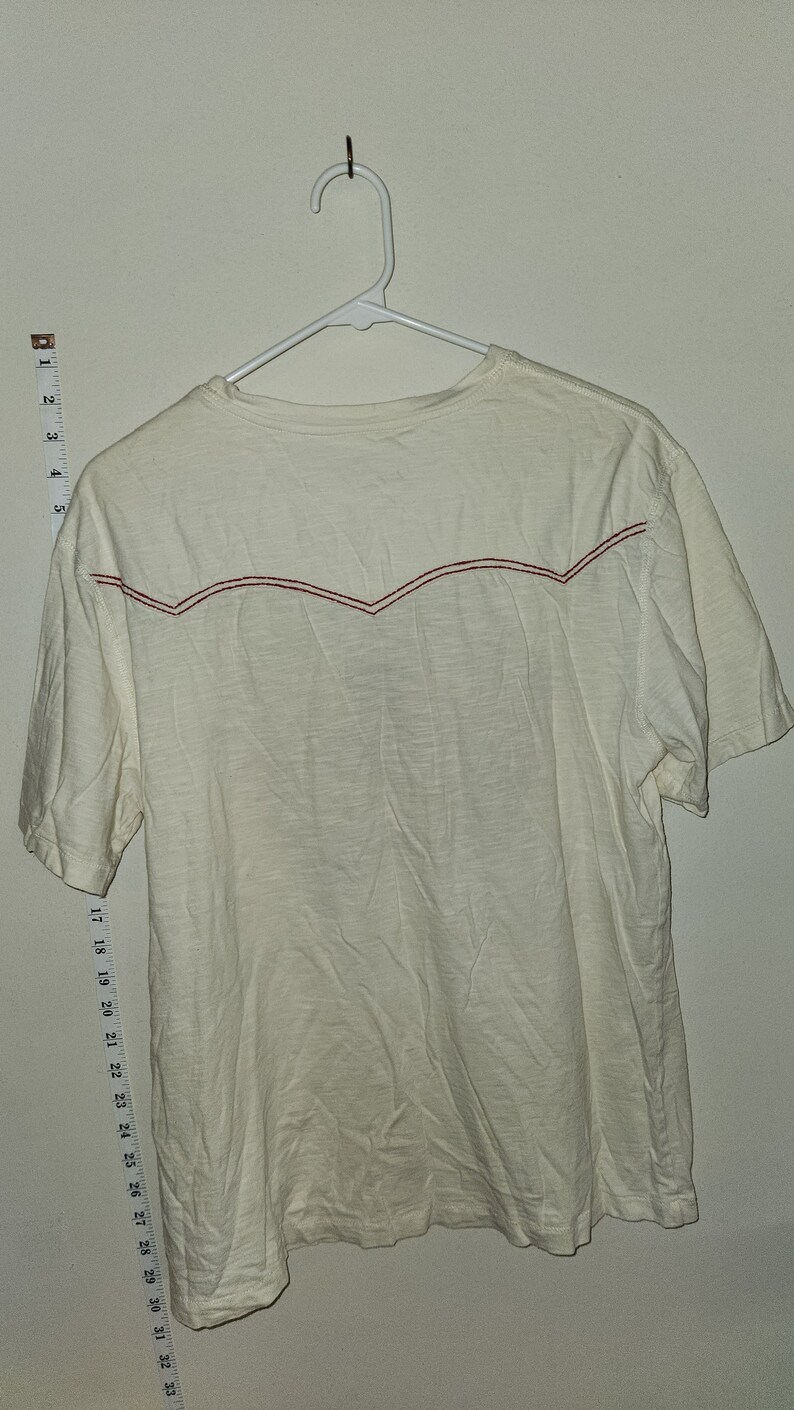Puede incluir: Una camiseta blanca de manga corta con un dise&ntilde;o bordado rojo en la espalda. La camiseta est&aacute; colgada en una percha blanca.