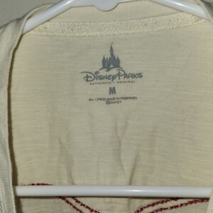 Puede incluir: Una camiseta blanca con un dise&ntilde;o bordado en rojo en el cuello. La camiseta tiene una etiqueta con el texto "Disney Parks Authentic Original" y una talla "M". La etiqueta tambi&eacute;n dice "RN 129855 Made in Philippines &copy;Disney".