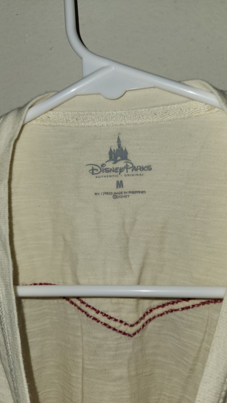 Puede incluir: Una camiseta blanca con un dise&ntilde;o bordado en rojo en el cuello. La camiseta tiene una etiqueta con el texto "Disney Parks Authentic Original" y una talla "M". La etiqueta tambi&eacute;n dice "RN 129855 Made in Philippines &copy;Disney".