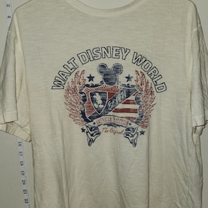 Puede incluir: Camiseta blanca con un gr&aacute;fico de estilo vintage del logotipo de Walt Disney World. El logotipo presenta a Mickey Mouse, estrellas y el texto "Walt Disney World" en un c&iacute;rculo. El texto "American Tradition" y "The Original" tambi&eacute;n est&aacute;n incluidos en el gr&aacute;fico.