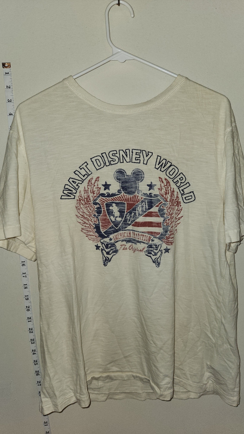Puede incluir: Camiseta blanca con un gr&aacute;fico de estilo vintage del logotipo de Walt Disney World. El logotipo presenta a Mickey Mouse, estrellas y el texto "Walt Disney World" en un c&iacute;rculo. El texto "American Tradition" y "The Original" tambi&eacute;n est&aacute;n incluidos en el gr&aacute;fico.