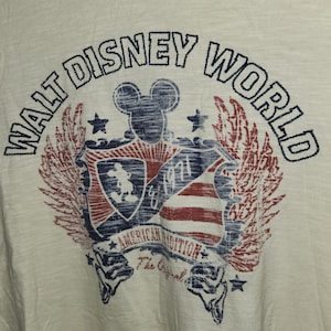 Puede incluir: Una camiseta blanca con un gr&aacute;fico de estilo vintage de Mickey Mouse y las palabras "Walt Disney World" en un esquema de colores azul y rojo. El gr&aacute;fico tambi&eacute;n incluye un escudo con un dise&ntilde;o de bandera estadounidense y el texto "American Tradition The Original".