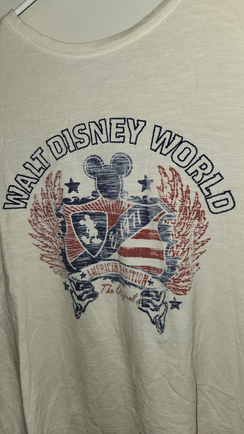 Puede incluir: Una camiseta blanca con un gr&aacute;fico de estilo vintage de Mickey Mouse y las palabras "Walt Disney World" en un esquema de colores azul y rojo. El gr&aacute;fico tambi&eacute;n incluye un escudo con un dise&ntilde;o de bandera estadounidense y el texto "American Tradition The Original".