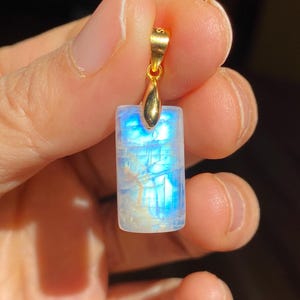 May include: A rectangular pendant with a gold bail, showcasing a luminous, iridescent moonstone. The stone displays a captivating play of blue and white light. The pendant is held in a hand against a dark background.
