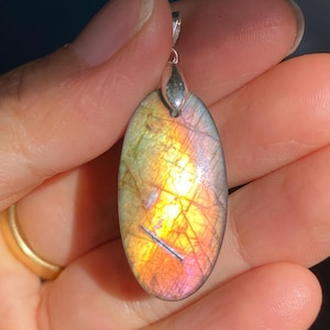 May include: Oval labradorite pendant with a silver bail. The gemstone exhibits a vibrant play of colour, with shades of orange, yellow, and purple. The pendant is held in a hand, showcasing the polished surface and natural patterns.