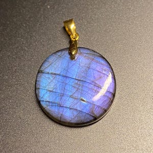 May include: A circular labradorite pendant featuring a polished surface with iridescent blue and purple flashes, characterised by natural internal fractures and inclusions, suspended from a simple gold-toned metal bail at the top centre.