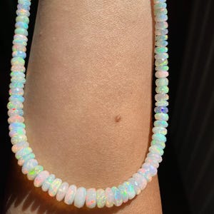 May include: A white opal bead necklace with iridescent flashes of colour. The beads are faceted and strung together, creating a delicate and eye-catching piece of jewellery. The necklace is shown on a person's arm.