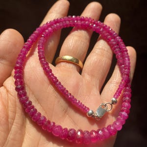 Op de afbeelding: Een ketting bestaande uit kleine, gefacetteerde, levendige roze-rode edelsteenkralen in een cirkelvormig patroon. De streng is voorzien van een gepolijste zilverkleurige metalen karabijnsluiting en een ringetje voor een veilige sluiting.