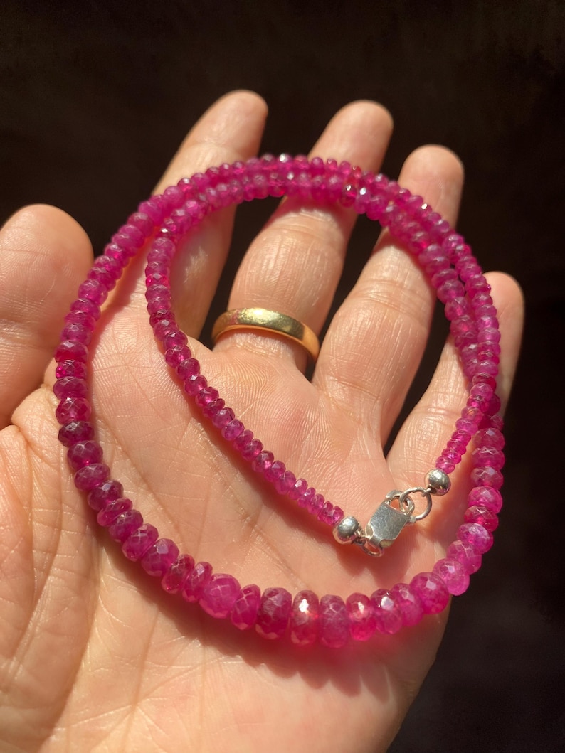 Op de afbeelding: Een ketting bestaande uit kleine, gefacetteerde, levendige roze-rode edelsteenkralen in een cirkelvormig patroon. De streng is voorzien van een gepolijste zilverkleurige metalen karabijnsluiting en een ringetje voor een veilige sluiting.