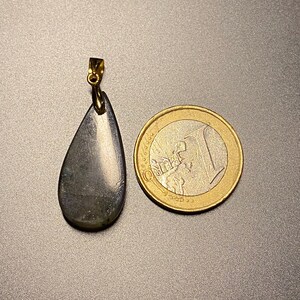 May include: A teardrop-shaped pendant with a gold-colored bail. The pendant is a dark, iridescent gray-green color. A one Euro coin is next to the pendant for size comparison. The pendant is likely a gemstone.