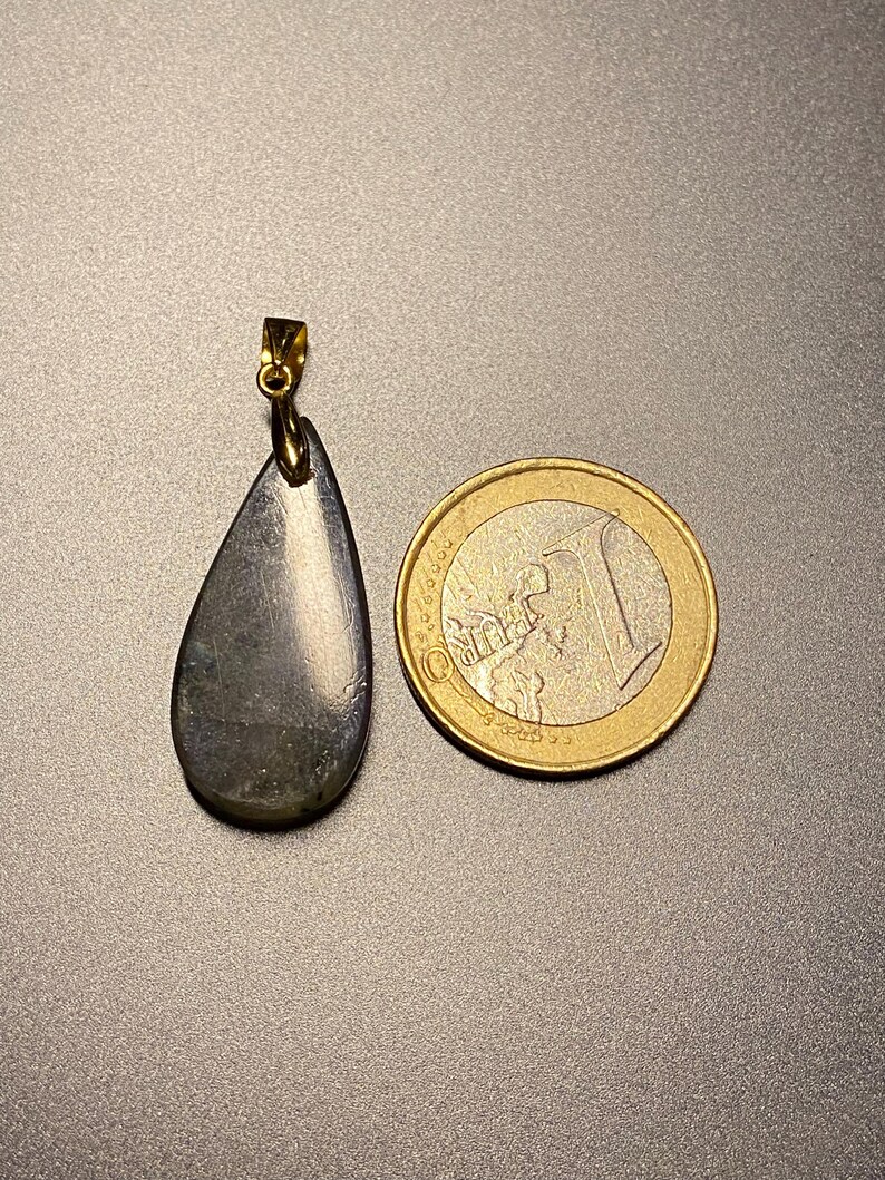 May include: A teardrop-shaped pendant with a gold-colored bail. The pendant is a dark, iridescent gray-green color. A one Euro coin is next to the pendant for size comparison. The pendant is likely a gemstone.