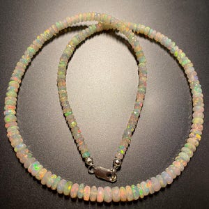 May include: A necklace made of small, iridescent opal beads. The beads are strung together, forming a circular shape. The necklace has a silver clasp. The opals display a range of colours, including white, green, orange, and pink.