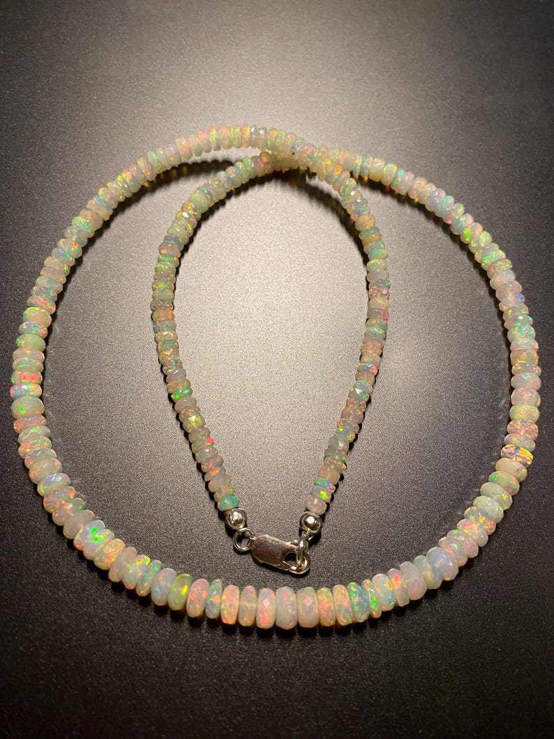 May include: A necklace made of small, iridescent opal beads. The beads are strung together, forming a circular shape. The necklace has a silver clasp. The opals display a range of colours, including white, green, orange, and pink.
