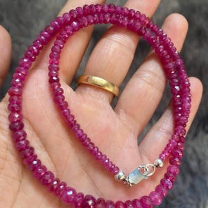 Op de afbeelding: Een ketting bestaande uit kleine, gefacetteerde, donkerroze robijnkralen gerangschikt in een cirkelvormig patroon. De streng is voorzien van een zilverkleurige karabijnsluiting en een ringetje die de uiteinden van de edelsteenkralenketting verbinden.
