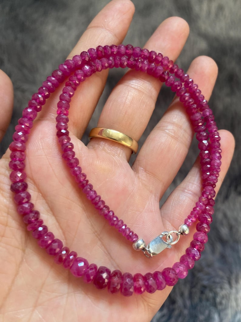 Op de afbeelding: Een ketting bestaande uit kleine, gefacetteerde, donkerroze robijnkralen gerangschikt in een cirkelvormig patroon. De streng is voorzien van een zilverkleurige karabijnsluiting en een ringetje die de uiteinden van de edelsteenkralenketting verbinden.