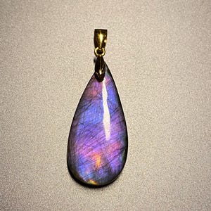 May include: A teardrop-shaped pendant with a gold-colored bail. The pendant is a polished gemstone, displaying a vibrant mix of purple, blue, and gold colors. The gemstone has a pearlescent sheen and is set against a neutral background.