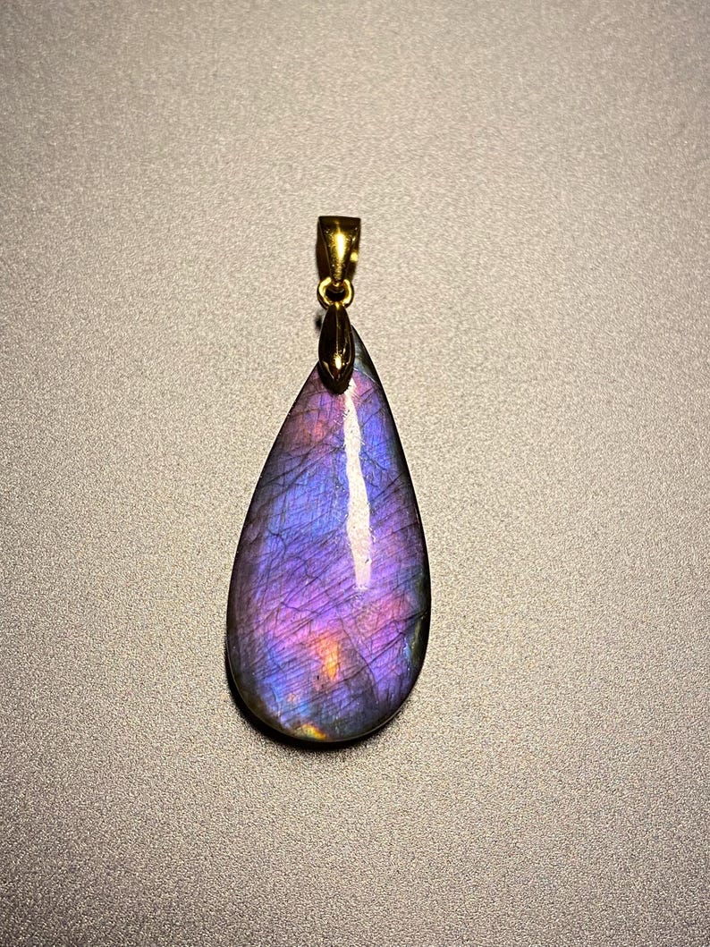 May include: A teardrop-shaped pendant with a gold-colored bail. The pendant is a polished gemstone, displaying a vibrant mix of purple, blue, and gold colors. The gemstone has a pearlescent sheen and is set against a neutral background.