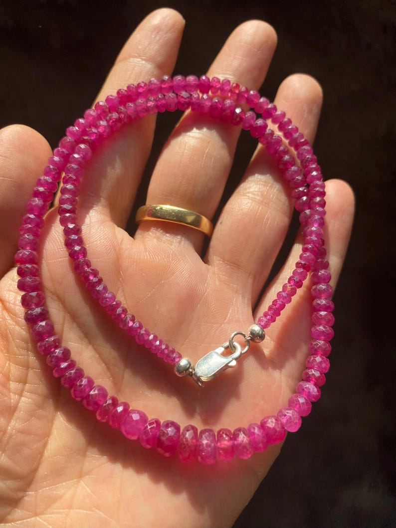 Op de afbeelding: Een ketting bestaande uit kleine, gefacetteerde, dieproze robijnkralen die aan elkaar zijn geregen. De streng is voorzien van een gepolijste, zilverkleurige karabijnsluiting aan de uiteinden voor een veilige sluiting van de kralen.