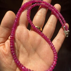 Op de afbeelding: Een ketting bestaande uit kleine, gefacetteerde, dieproze robijnkralen die aan elkaar zijn geregen. De streng heeft aan &eacute;&eacute;n uiteinde een metalen zilverkleurige sluiting die de cirkelvormige opstelling van gepolijste edelsteenkralen vasthoudt.