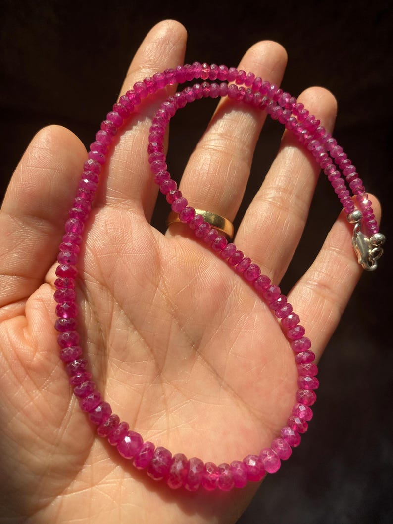 Op de afbeelding: Een ketting bestaande uit kleine, gefacetteerde, dieproze robijnkralen die aan elkaar zijn geregen. De streng heeft aan &eacute;&eacute;n uiteinde een metalen zilverkleurige sluiting die de cirkelvormige opstelling van gepolijste edelsteenkralen vasthoudt.
