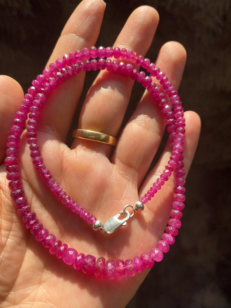 Op de afbeelding: Een ketting bestaande uit kleine, gefacetteerde, levendige magenta-roze edelsteenkralen. De streng is voorzien van een gepolijste zilverkleurige karabijnsluiting aan de uiteinden, wat zorgt voor een veilige sluiting van de cirkelvormige kralen.