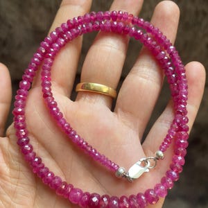 Op de afbeelding: Een streng van gefacetteerde, dieproze robijnkralen gerangschikt in een cirkelvormig patroon. De ketting bestaat uit kleine, uniforme, doorschijnende edelsteenkralen met een gepolijste afwerking en een zilverkleurige metalen karabijnsluiting.