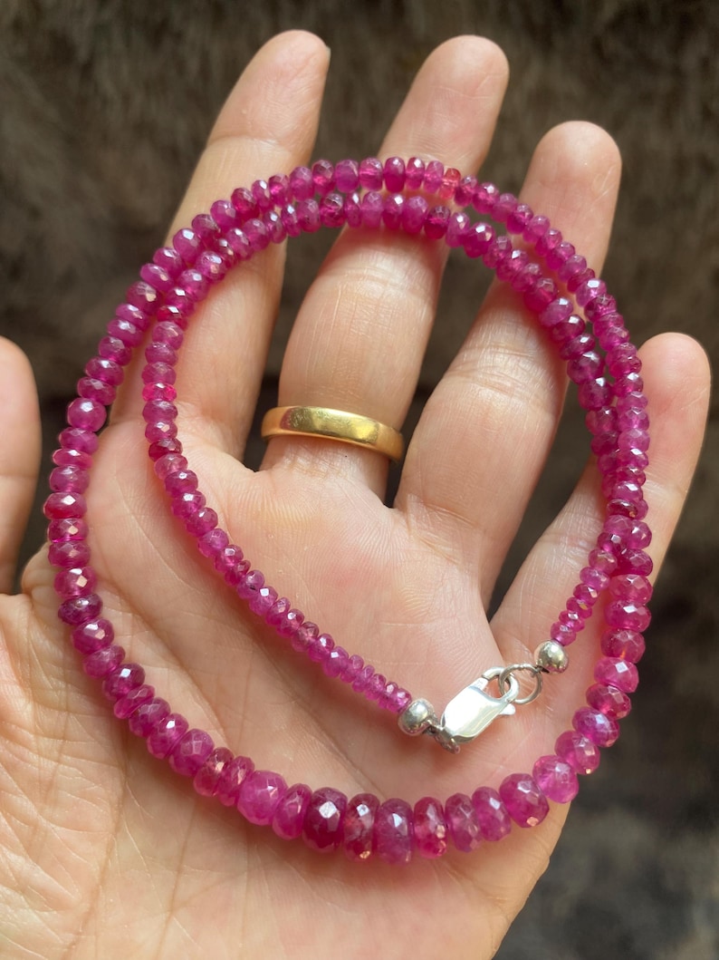 Op de afbeelding: Een streng van gefacetteerde, dieproze robijnkralen gerangschikt in een cirkelvormig patroon. De ketting bestaat uit kleine, uniforme, doorschijnende edelsteenkralen met een gepolijste afwerking en een zilverkleurige metalen karabijnsluiting.