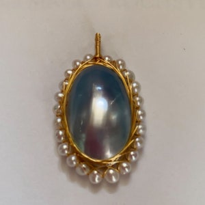 Peut inclure: Pendentif en or avec une pierre ovale irisée, encadrée de petites perles et de fil d'or. La pierre reflète la lumière bleue et argentée. Le pendentif a une bélière pour une chaîne. Design élégant et détaillé.