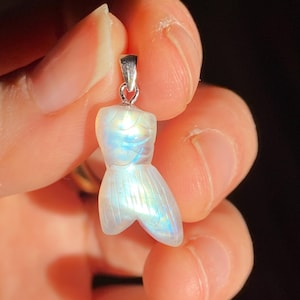 以下が含まれることがあります： 銀色のベイルが付いた、真珠光沢のある虹色の人魚の尾のペンダント。ペンダントは半透明の石から彫られており、青、緑、黄色の色合いを示しています。人魚の尾のデザインは詳細で滑らかです。