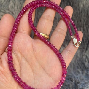 Op de afbeelding: Een ketting bestaande uit kleine, gefacetteerde, diep roze-rode edelsteenkralen. Het snoer is voorzien van een gepolijste, zilverkleurige metalen karabijnsluiting aan het uiteinde, wat zorgt voor een veilige sluiting van het ronde kralensieraad.