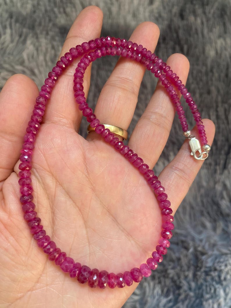 Op de afbeelding: Een ketting bestaande uit kleine, gefacetteerde, diep roze-rode edelsteenkralen. Het snoer is voorzien van een gepolijste, zilverkleurige metalen karabijnsluiting aan het uiteinde, wat zorgt voor een veilige sluiting van het ronde kralensieraad.