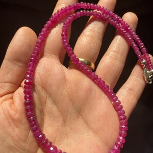 Op de afbeelding: Een ketting bestaande uit kleine, gefacetteerde, dieproze-rode edelsteenkralen. De kralen zijn geregen aan een enkele streng en nemen geleidelijk in grootte toe naar het midden. De ketting heeft een zilverkleurige metalen karabijnsluiting.