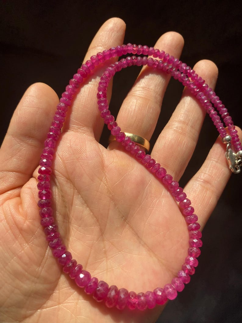 Op de afbeelding: Een ketting bestaande uit kleine, gefacetteerde, dieproze-rode edelsteenkralen. De kralen zijn geregen aan een enkele streng en nemen geleidelijk in grootte toe naar het midden. De ketting heeft een zilverkleurige metalen karabijnsluiting.