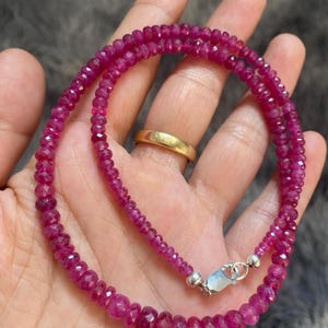 Op de afbeelding: Een ketting bestaande uit kleine, gefacetteerde, diep roze-rode robijnkralen gerangschikt in een cirkelvormig patroon. De streng is voorzien van een zilverkleurige karabijnsluiting en kleine zilverkleurige kraaltjes aan de uiteinden.