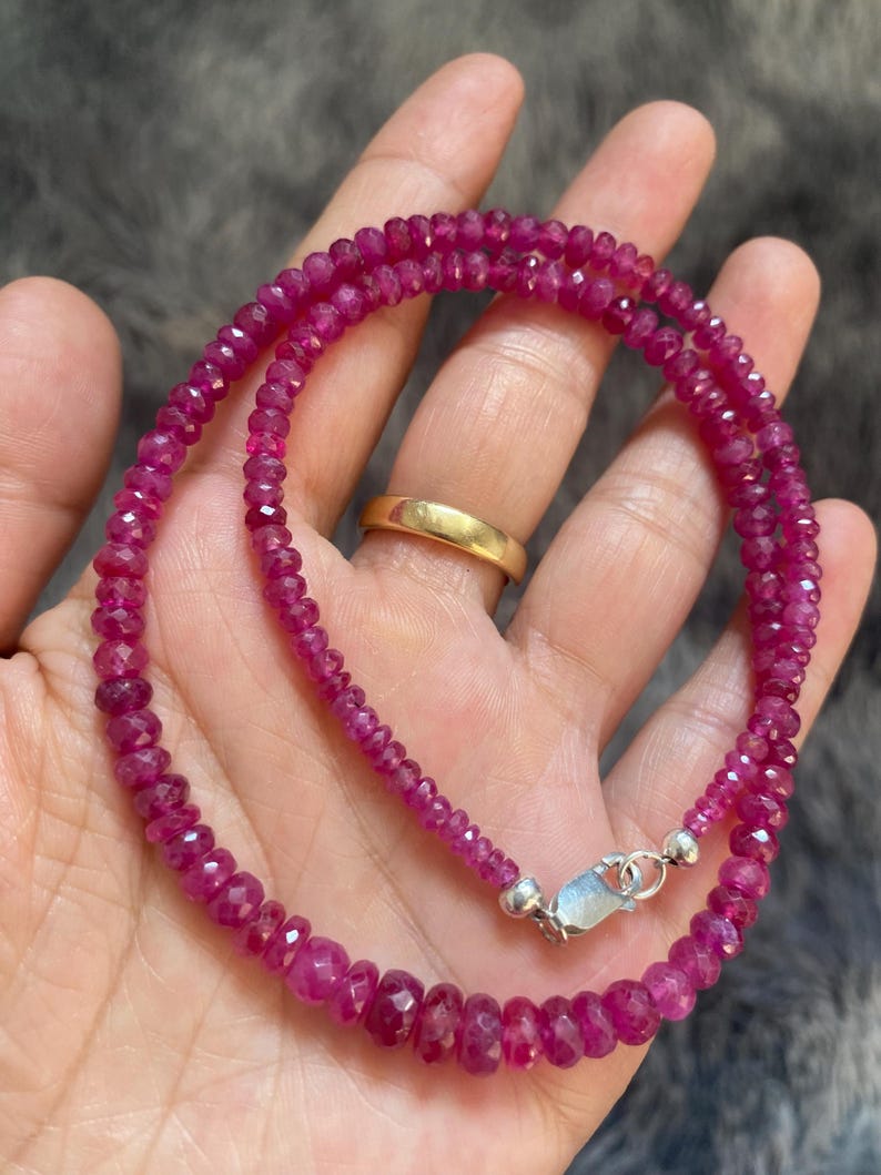 Op de afbeelding: Een ketting bestaande uit kleine, gefacetteerde, diep roze-rode robijnkralen gerangschikt in een cirkelvormig patroon. De streng is voorzien van een zilverkleurige karabijnsluiting en kleine zilverkleurige kraaltjes aan de uiteinden.