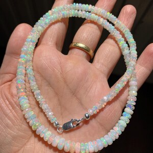 May include: A necklace made of small, iridescent opal beads. The beads are strung together, creating a shimmering effect. The necklace is secured with a silver clasp. The opal beads display a range of colours, including white, blue, and hints of pink.