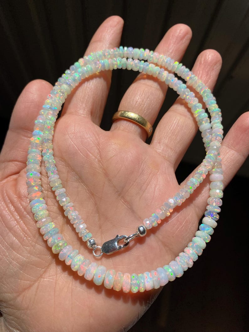 May include: A necklace made of small, iridescent opal beads. The beads are strung together, creating a shimmering effect. The necklace is secured with a silver clasp. The opal beads display a range of colours, including white, blue, and hints of pink.