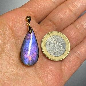 May include: A teardrop-shaped pendant with a vibrant purple and blue iridescent gemstone, set in a gold-colored metal. The pendant is next to a Euro coin for size comparison. The gemstone displays a play of color.