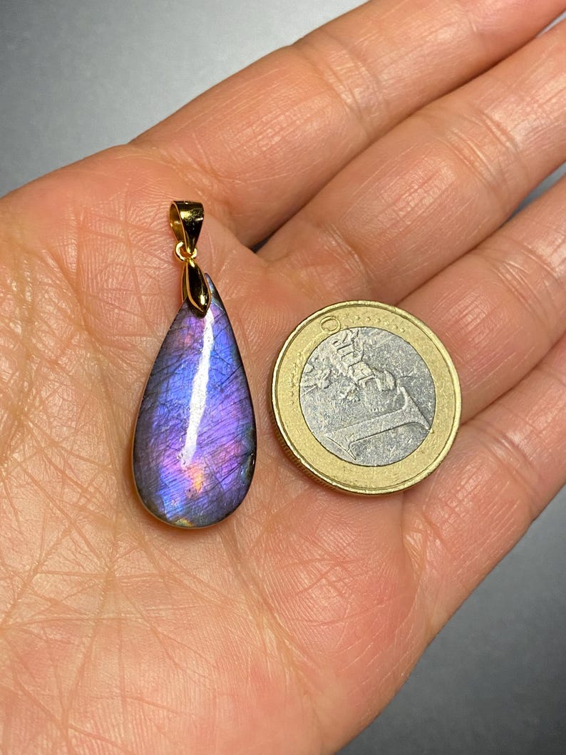 May include: A teardrop-shaped pendant with a vibrant purple and blue iridescent gemstone, set in a gold-colored metal. The pendant is next to a Euro coin for size comparison. The gemstone displays a play of color.