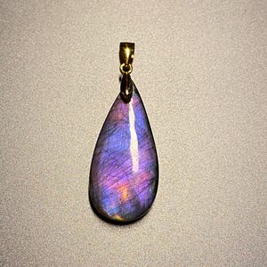 May include: A teardrop-shaped pendant with a gold-colored bail. The pendant is a polished stone with a mix of purple, blue, and orange hues. The stone has a reflective surface and is set against a gray background.