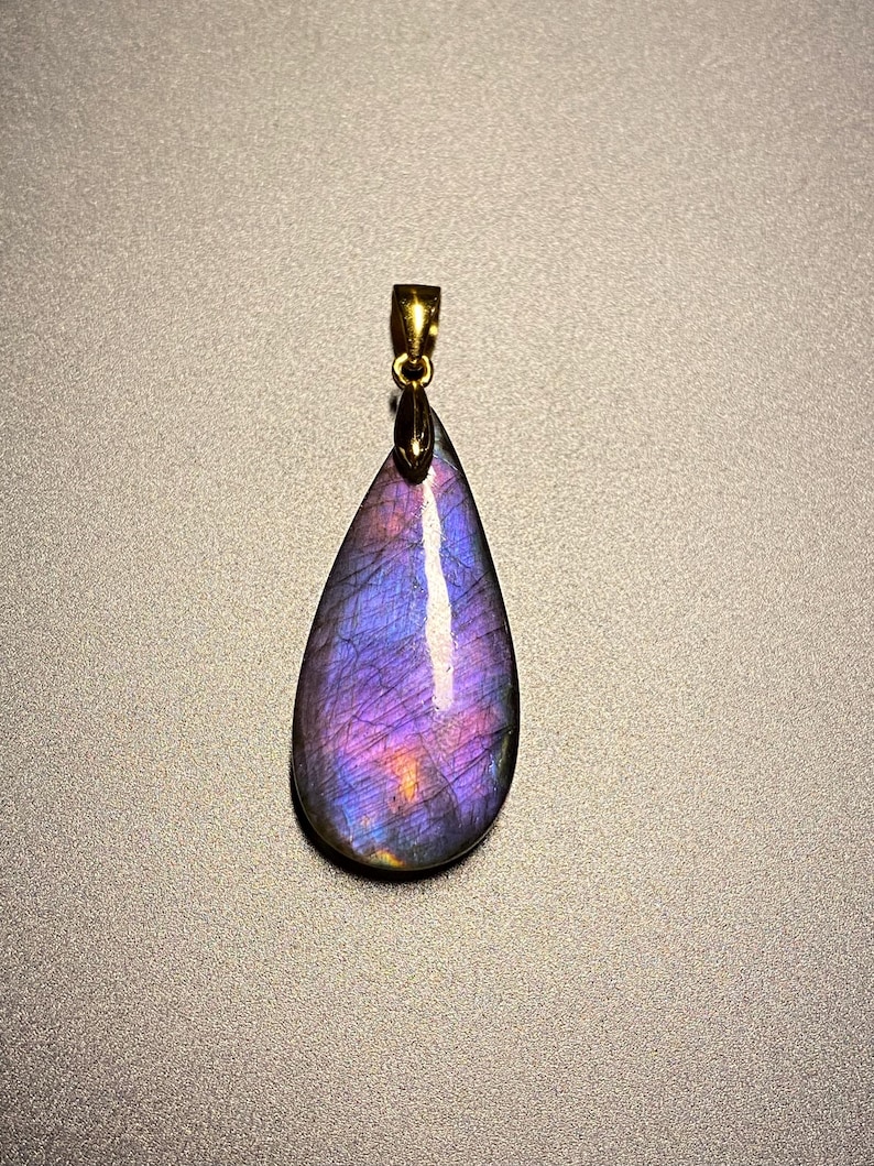 May include: A teardrop-shaped pendant with a gold-colored bail. The pendant is a polished stone with a mix of purple, blue, and orange hues. The stone has a reflective surface and is set against a gray background.