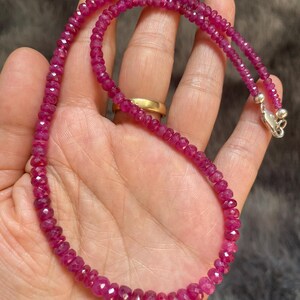 Op de afbeelding: Een ketting bestaande uit kleine, gefacetteerde, diep roze-rode edelsteenkralen die aan elkaar zijn geregen. De streng is voorzien van een gepolijste zilverkleurige metalen karabijnsluiting aan het uiteinde voor een veilige sluiting.