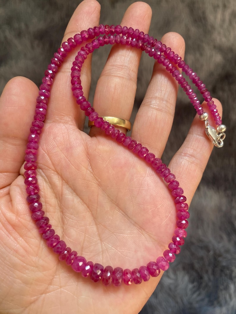 Op de afbeelding: Een ketting bestaande uit kleine, gefacetteerde, diep roze-rode edelsteenkralen die aan elkaar zijn geregen. De streng is voorzien van een gepolijste zilverkleurige metalen karabijnsluiting aan het uiteinde voor een veilige sluiting.