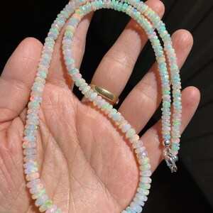 May include: A necklace made of small, iridescent opal beads. The beads show a variety of colours, including white, orange, green, and blue. The necklace is fastened with a silver clasp. The jewellery is displayed against a dark background.