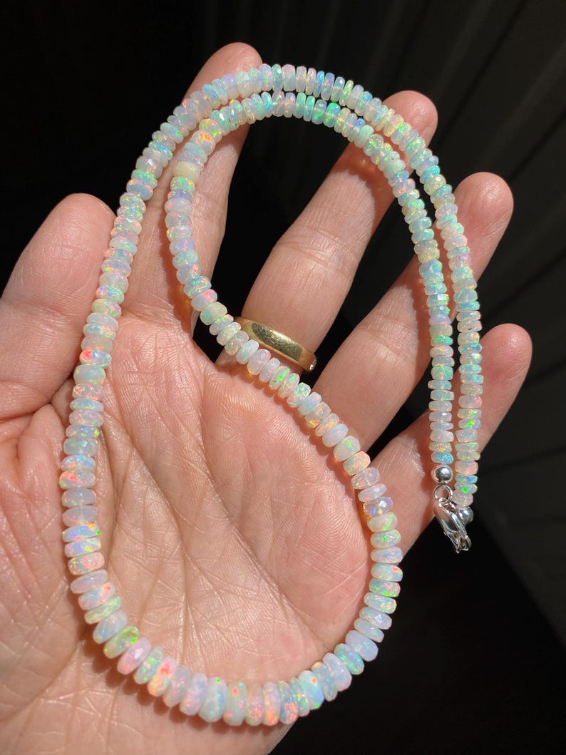 May include: A necklace made of small, iridescent opal beads. The beads show a variety of colours, including white, orange, green, and blue. The necklace is fastened with a silver clasp. The jewellery is displayed against a dark background.