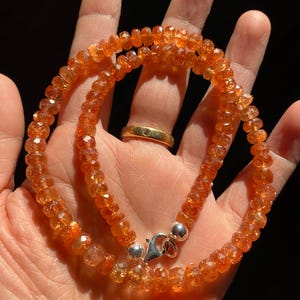 Può includere: Collana composta da piccole gemme arancioni sfaccettate. Le perline sono infilate, formando un doppio filo assicurato con una chiusura argentata. La collana è tenuta in una mano, con un anello d'oro visibile.