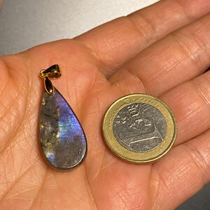 May include: A teardrop-shaped labradorite pendant with a gold-colored bail. The pendant displays iridescent blue, purple, and gray hues. A one Euro coin is next to the pendant for size comparison.
