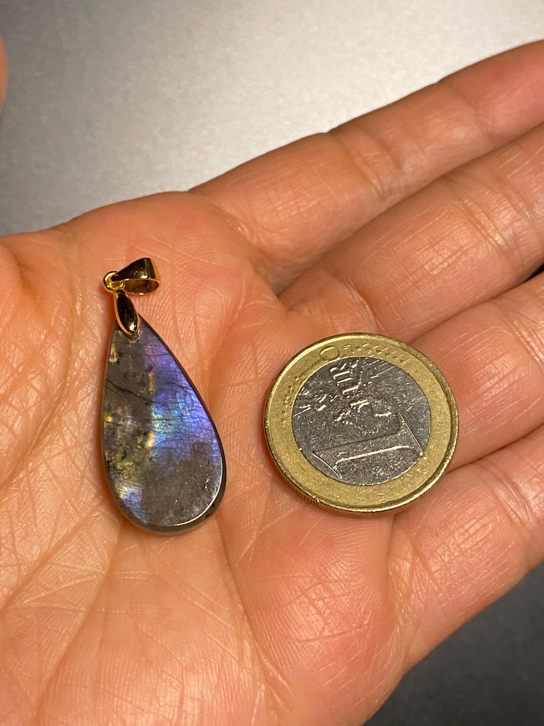May include: A teardrop-shaped labradorite pendant with a gold-colored bail. The pendant displays iridescent blue, purple, and gray hues. A one Euro coin is next to the pendant for size comparison.