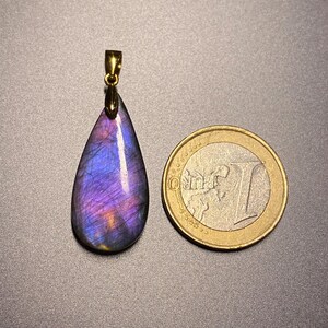 May include: A teardrop-shaped pendant with a gold-colored bail, showcasing a vibrant display of purple and blue hues. The pendant is next to a Euro coin for size comparison. The gemstone pendant is a unique piece of jewelry.