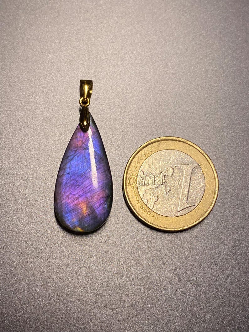 May include: A teardrop-shaped pendant with a gold-colored bail, showcasing a vibrant display of purple and blue hues. The pendant is next to a Euro coin for size comparison. The gemstone pendant is a unique piece of jewelry.